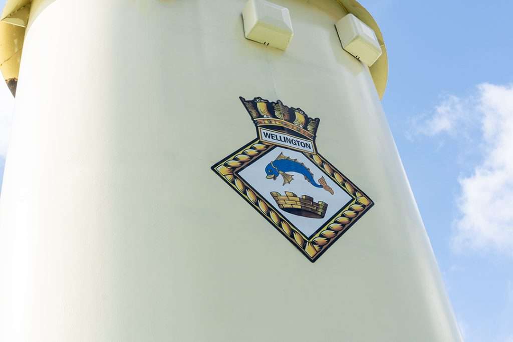HMS Wellington crest emblem displayed on a pale yellow funnel against a blue sky