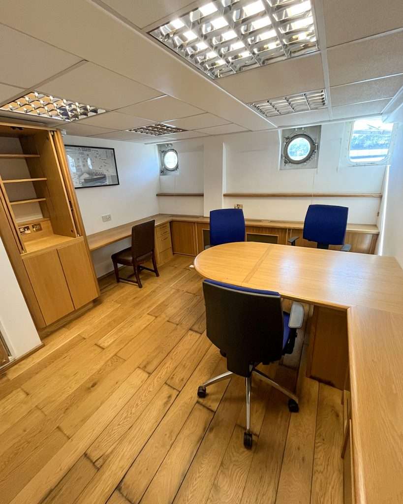 Office space on board HMS Wellington with wooden flooring, meeting table and blue chairs used for workspace hire